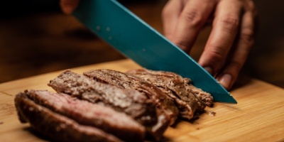 A person slices a steak with a knife on a wooden chopping board. Photo by Los Muertos Crew via Pexels.
