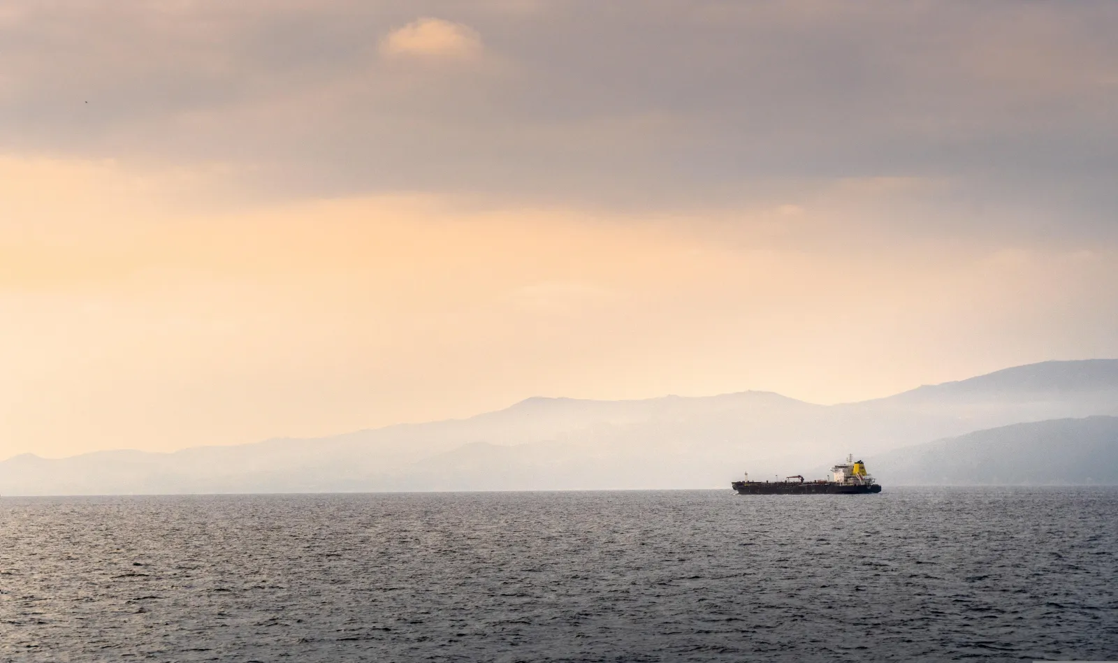 A photo of a lone cargo ship in the distance with water in the foreground and misty mountainous landscape behind it. Photo by rbardy24 via Pixabay.