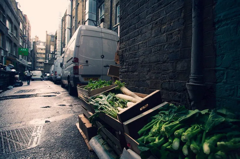 A large white van is parked in an alleyway next to boxes of vegetables