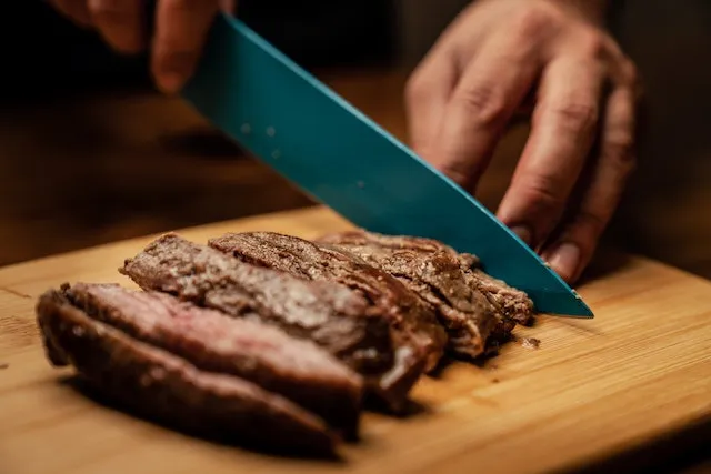 A person slices a steak with a knife on a wooden chopping board. Photo by Los Muertos Crew via Pexels.