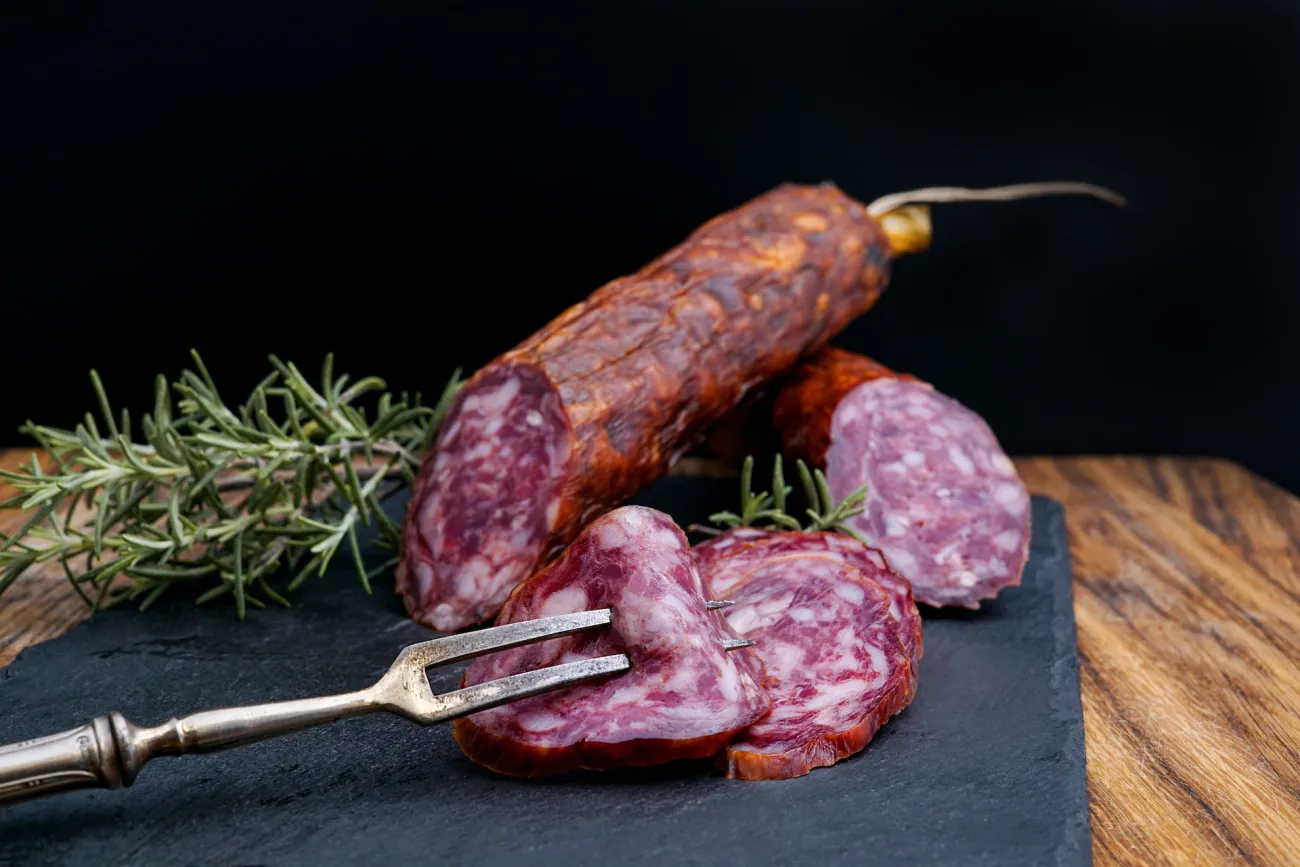 Salami sausage on black chopping board with rosemary and meat fork. Image by Wesual Click via Unsplash.