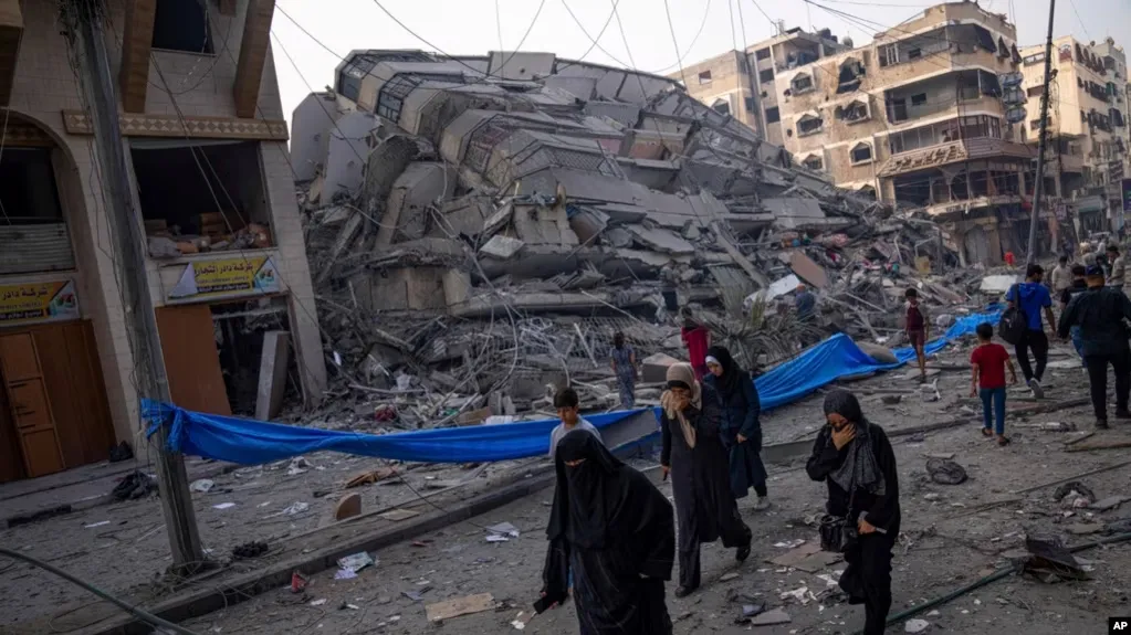 Palestinians walk by the rubble of a building after it was hit by an Israeli airstrike, in Gaza City, Photo via VOA News