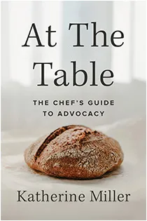 The cover of At the Table featuring a loaf of bread on a white tablecloth