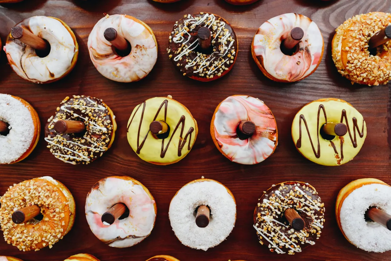 Frosted donuts hang on wooden pegs at a dessert stand. Photo by Anastasiia Chepinska via UnSplash.