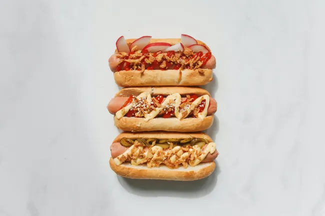 Image of three hot dogs with various toppings on white surface. Photo by Polina Tankilevitch via Pexels.