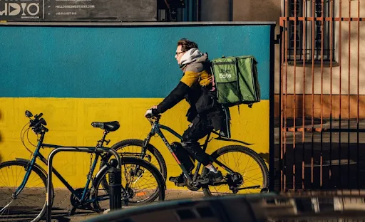 Uber Eats delivery person on an ebike rides past a wall painted like the Ukrainian flag. Photo by Ammy Singh via Pexels.
