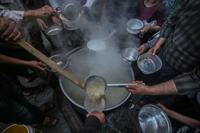 Photo of food being laddled from a large pot, surrounded by hands holding empty bowls in Gaza. Image by Yousef Masoud via Pixabay.