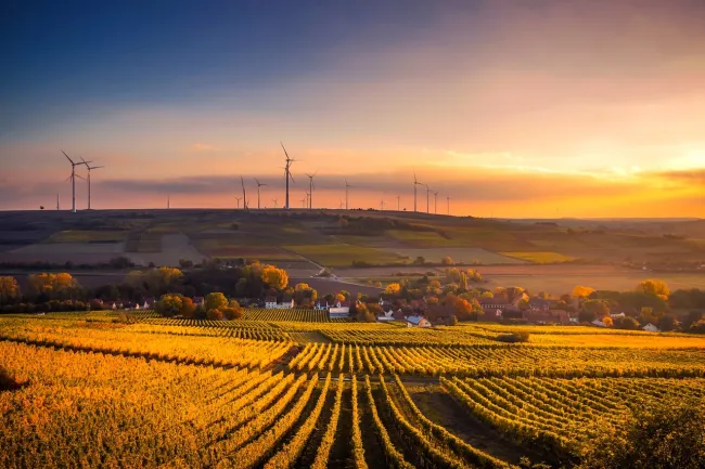A farm at sunset with wind turbines in the background. Photo by Pixabay from Pexels