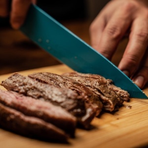 A person slices a steak with a knife on a wooden chopping board. Photo by Los Muertos Crew via Pexels.