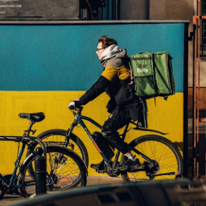 Uber Eats delivery person on an ebike rides past a wall painted like the Ukrainian flag. Photo by Ammy Singh via Pexels.